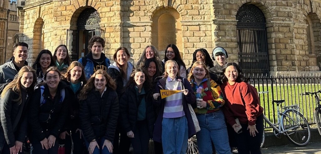 A group of about 15 students stand together on a sunny winter day outside the Bodleian Library in Oxford. One student is holding a small yellow PLU banner.