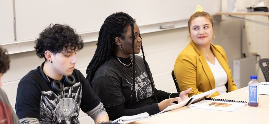 Students in and IHON class sit next to each other at a table in a small classroom. Books and notebooks are in front of them. One student is speaking while another looks on.