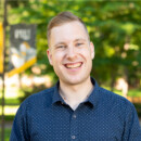 Jeffrey Roberts smiles, looking at the camera. He is wearing a blue button up shirt and behind him is the PLU campus on a sunny day.