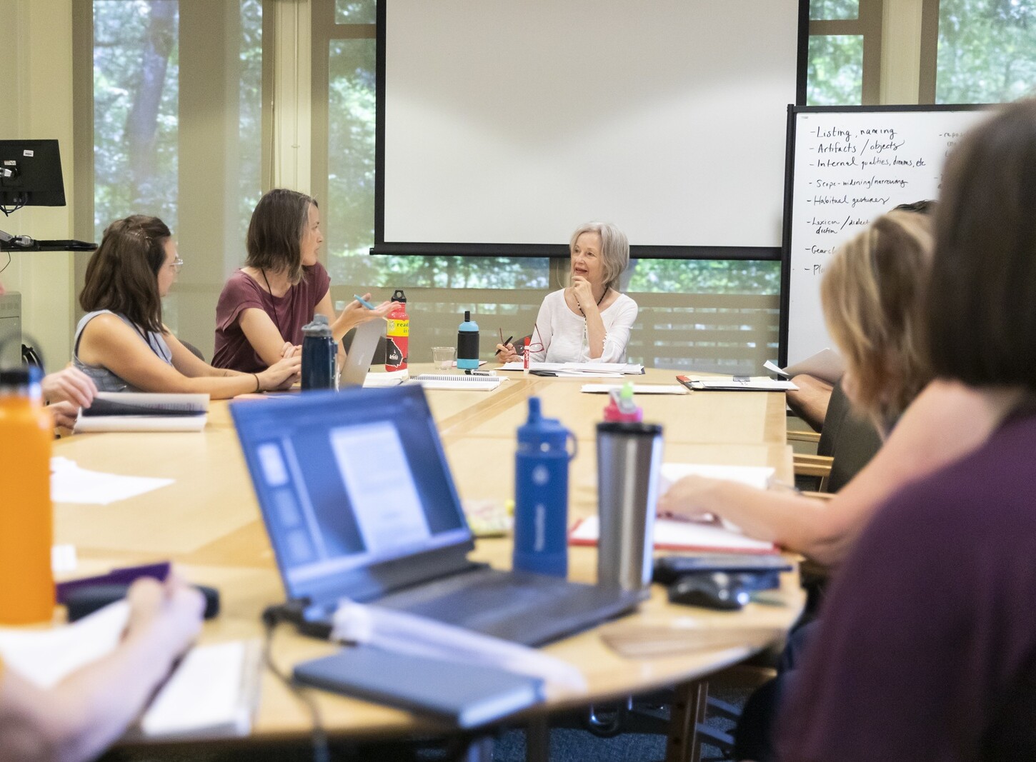 MFA students sit at an oblong table with laptops, notebooks, pens and water bottles in front of them. They are in discussion with the faculty at the head of the table.