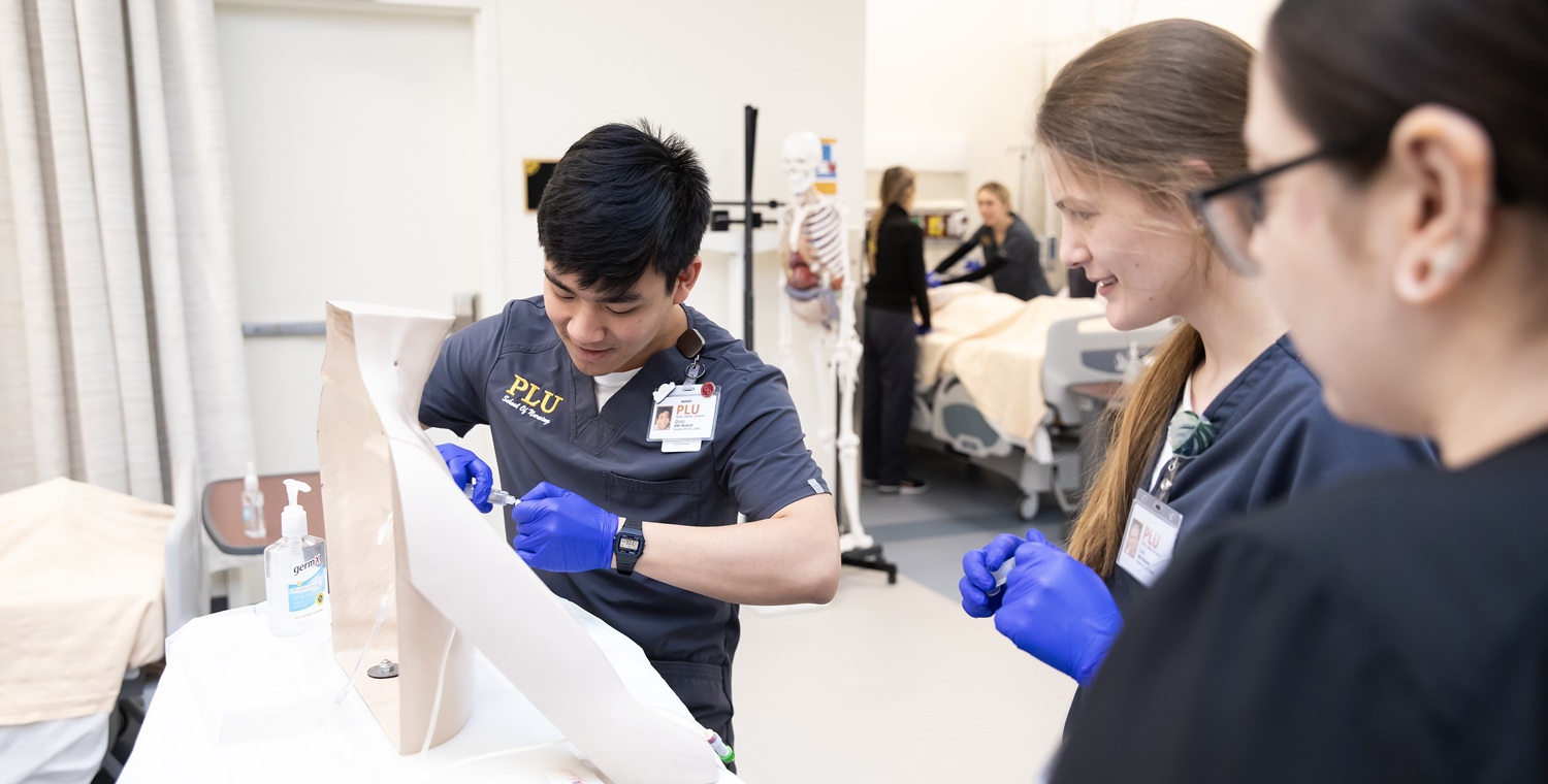 Three PLU Nursing students wearing dark blue PLU Nursing School scrubs and latex gloves practice placing central lines in the Nursing Simulation Center.