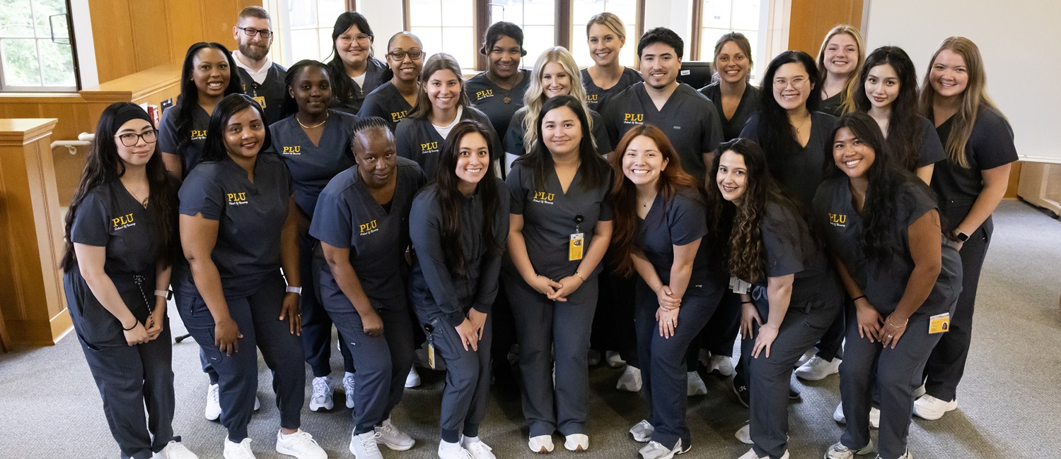 Web Banner - MSN Group 2025 The 2025 cohort of MSN students stand together for a group shot, smiling at the camera. There are about 20 students and all are wearing grey-blue scrubs that have 'PLU School of Nursing' embroidered on them.