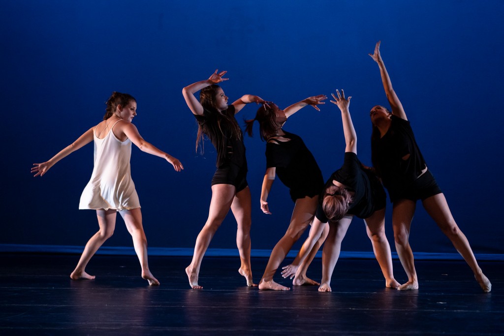 One dancer wearing white dances with four other dancers in black on a dark blue background.