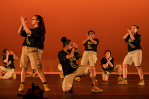PLU's step dance group, Lute Nation, poses with their arms crossed in front of their faces with an orange background behind the.