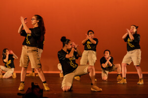 PLU's step dance group, Lute Nation, poses with their arms crossed in front of their faces with an orange background behind the.