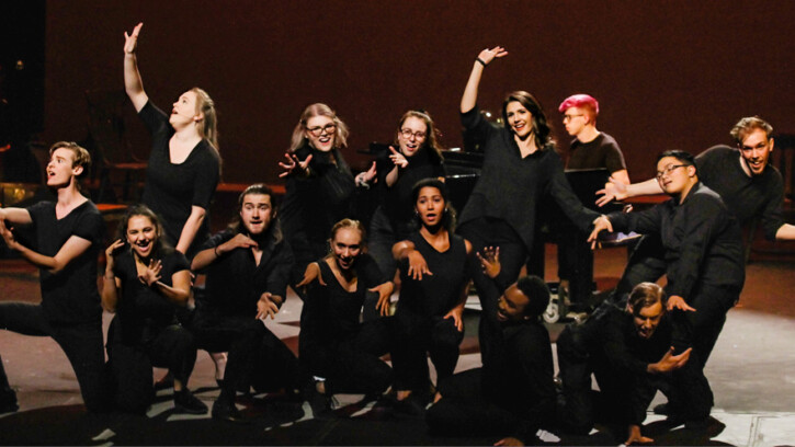 Musical theatre students dressed in black clump together onstage, some kneeling and some standing, with arms outstretched.