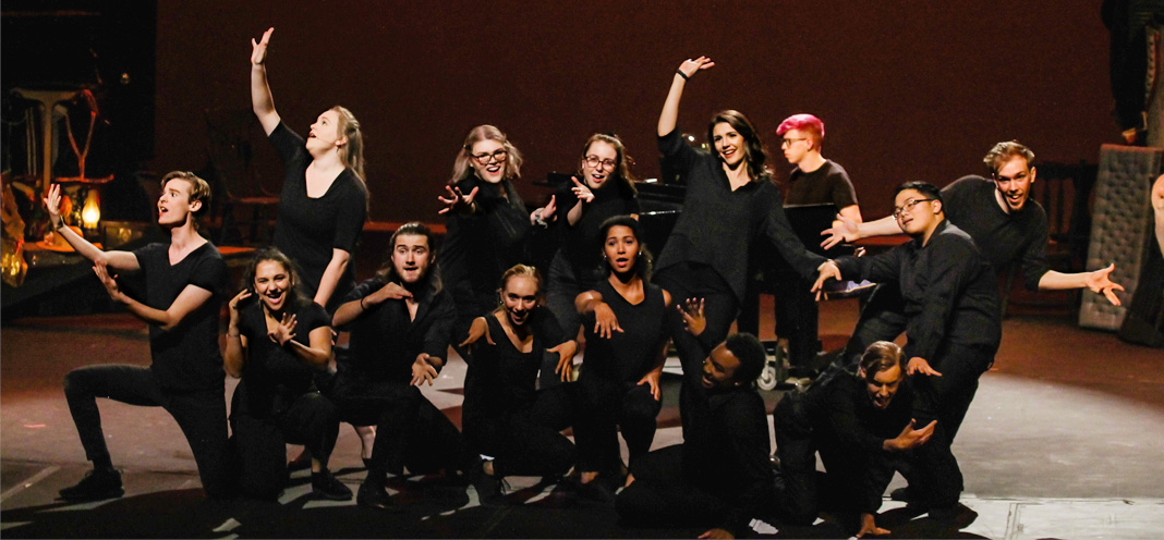 Musical theatre students dressed in black clump together onstage, some kneeling and some standing, with arms outstretched.
