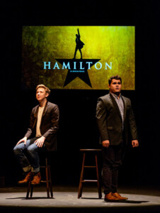 Two men sing in front of a backdrop that says "HAMILTON." One sits on a stool and the other stands.