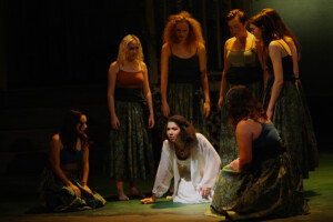 A group of female chorus members dressed in green surround Medea dressed in white.