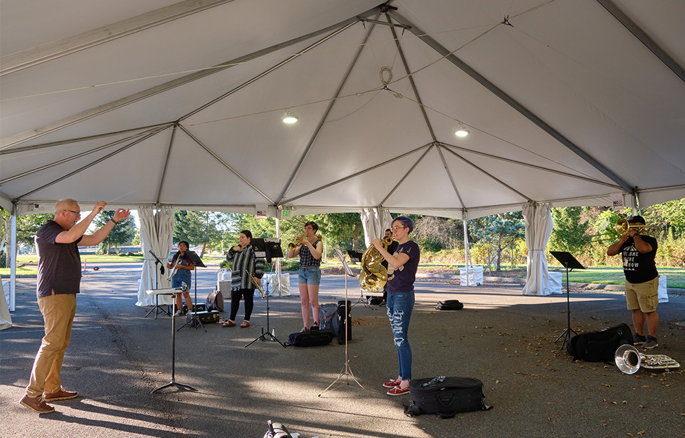 Students rehearse on brass instruments outdoors in a tent set up for safety during COVID-19 pandemic. Dr. Edwin Powell conducts.