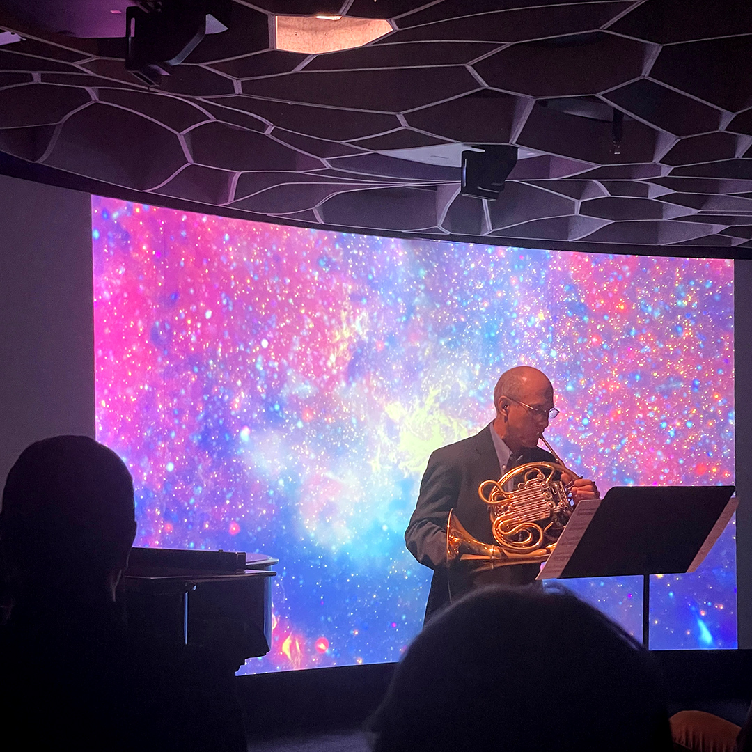 Mark Robbins, Associate Principal Horn of the Seattle Symphony, performing the premiere of Pale Blue Dot in front of a multicolored speckled galaxy