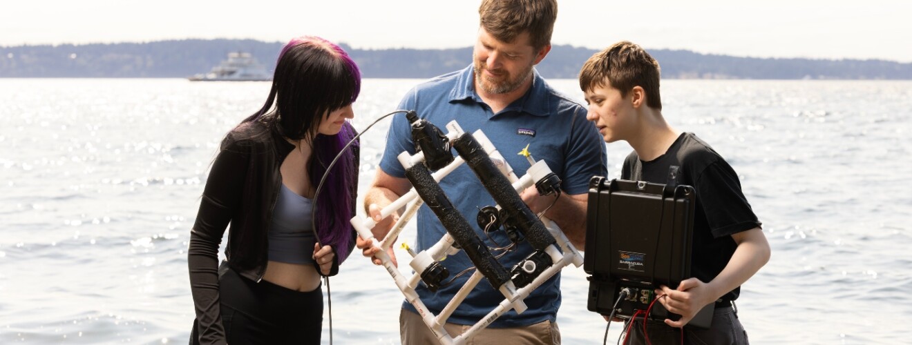 professor and two students looking at a drone on the beach.