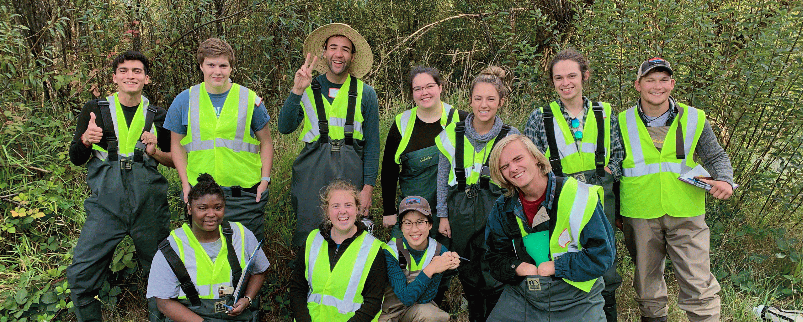 PLU students pose for a photo at Clover Creek, all wearing yellow construction vests over raingear.