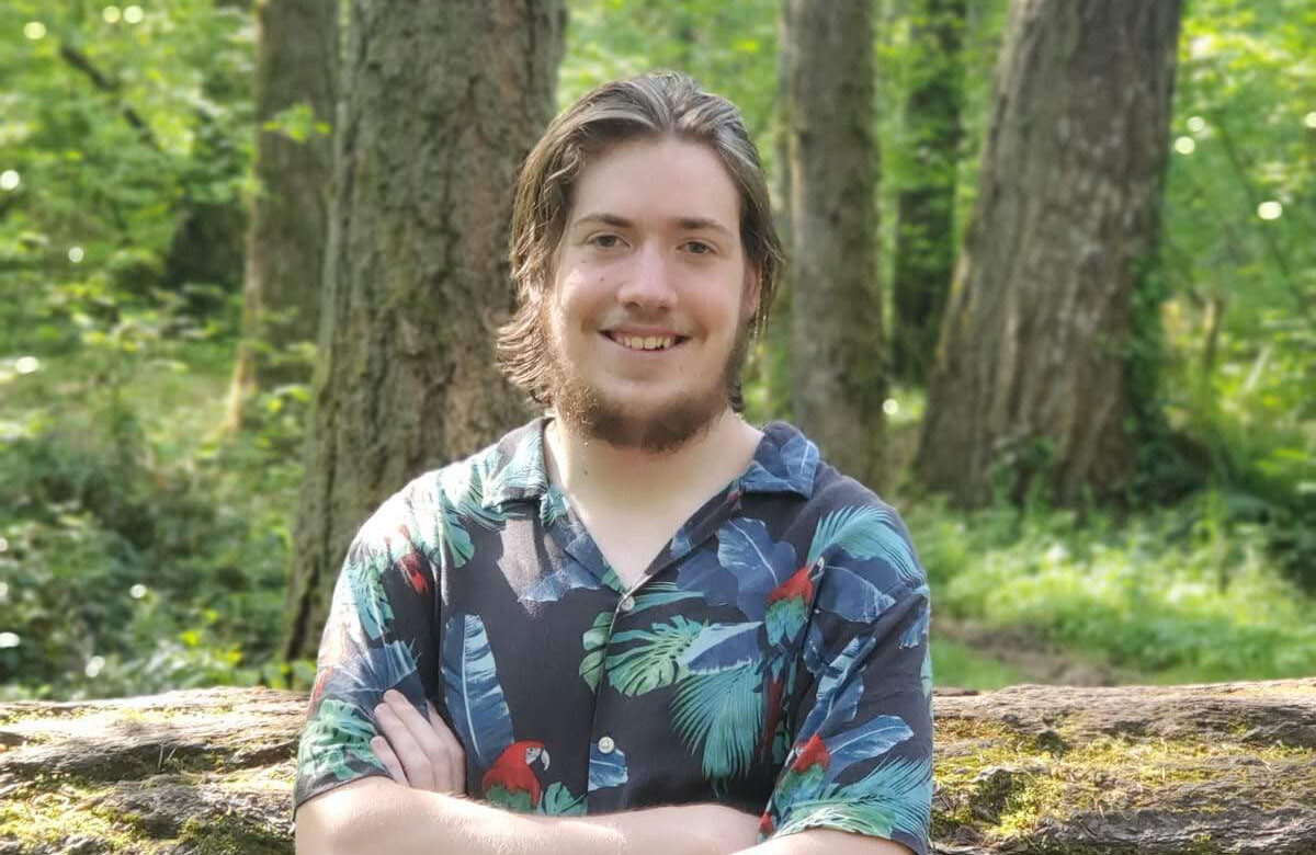 Daniel Hachet '20 stands with his arms folded and a forest behind him