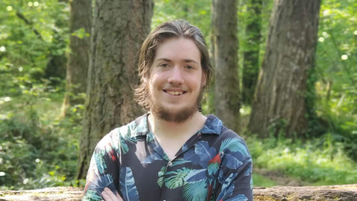 Daniel Hachet '20 stands with his arms folded and a forest behind him