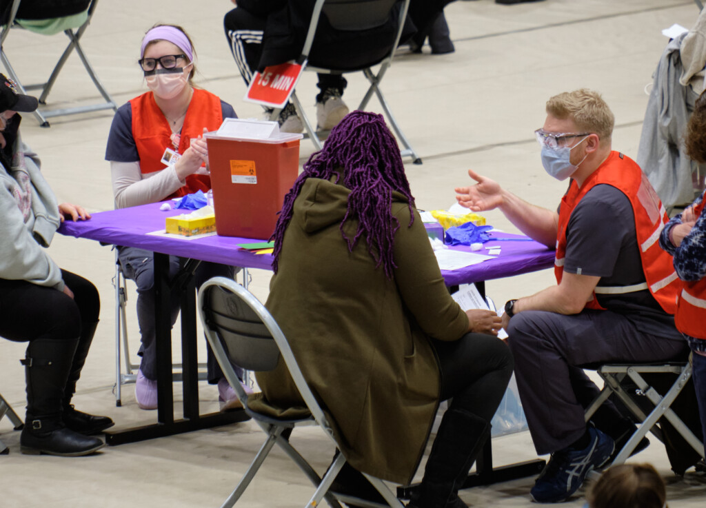 two PLU students, both wearing red vests, nursing scrubs, and facemasks work with patients at the COVID-19 vaccine clinic at PLU.