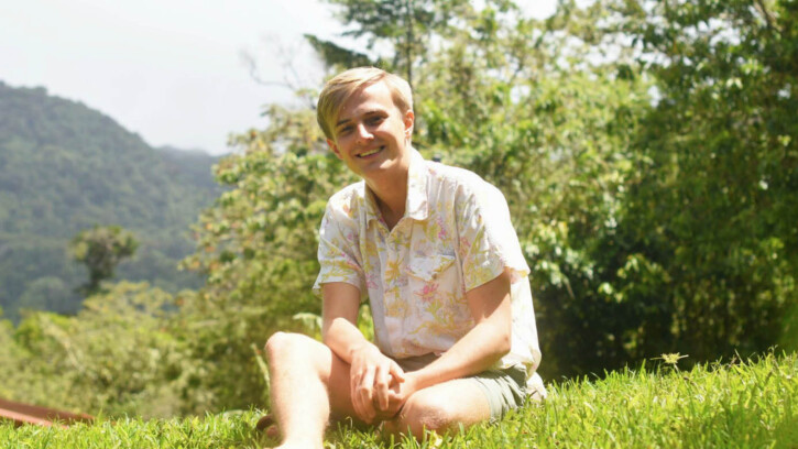 John sits cross-legged in a grass field on a sunny day with rolling hills behind him.