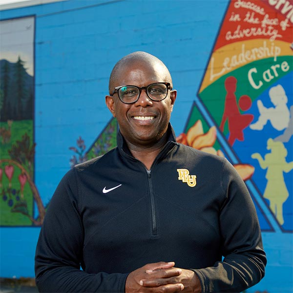 George Zeno stands smiling on front of of a brightly colored mural on Garfield Street near PLU.