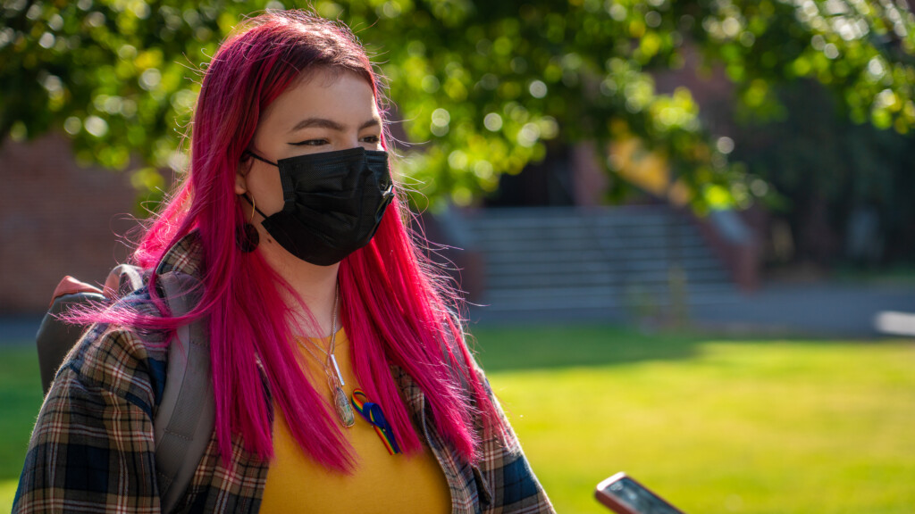 Student Ceci Omri ’24 wearing a black mask. She has long purple hair and is wearing a flannel longsleeve shirt.
