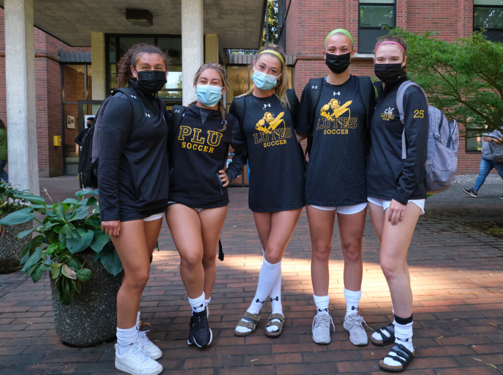 Five of friends wearing matching black long sleeve shirts from the women's soccer team pose for a photo together outside their residence hall.