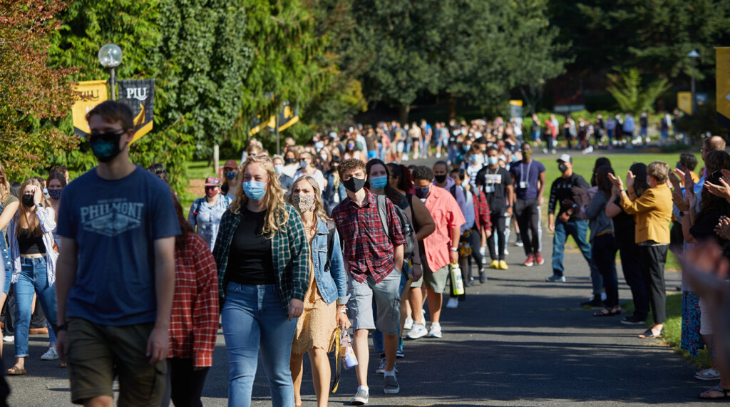 return-6 a wide photo of more than 100 students walking on the path next to Foss Field, on their way to convocation.