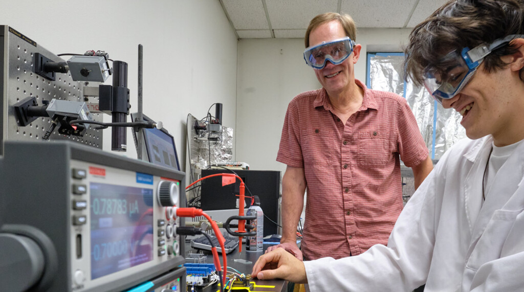 Professor Waldo and a student, both wearing protective goggles, share a laugh in a chemistry lab while working