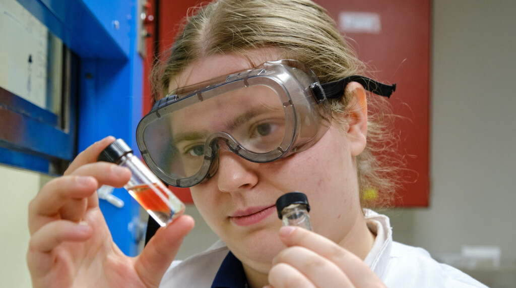 close of photo of a student in lab goggles looking at red substance in a small vile.