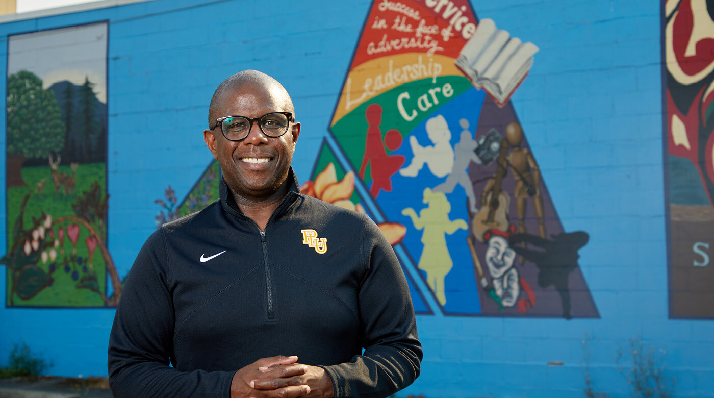 George Zeno stands smiling on front of of a brightly colored mural on Garfield Street near PLU.