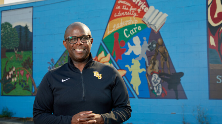 George Zeno stands smiling on front of of a brightly colored mural on Garfield Street near PLU.