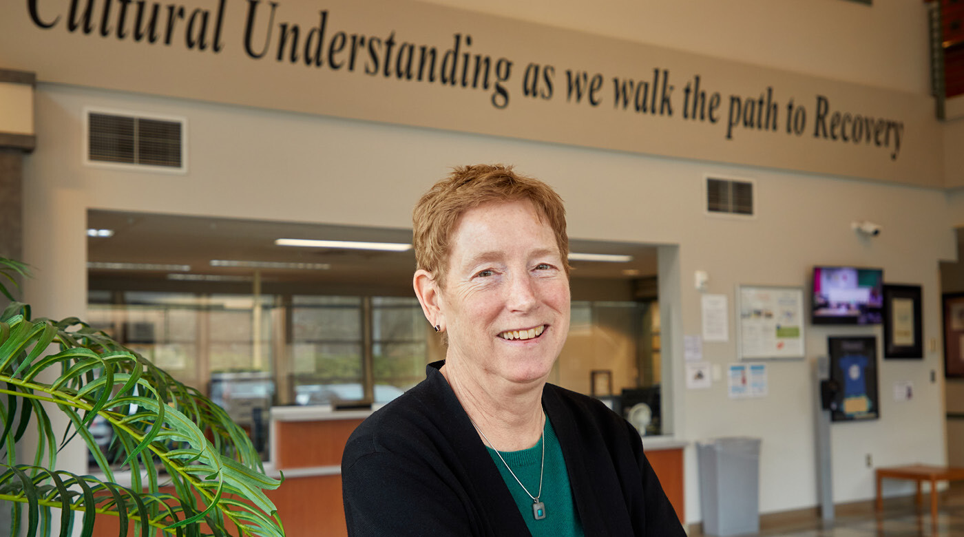 Teri Card smiles into the camera in the lobby of her office building. Above her head a quote painted on the wall reads "Cultural understanding as we walk the path to Recovery"