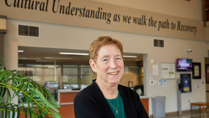 Teri Card smiles into the camera in the lobby of her office building. Above her head a quote painted on the wall reads "Cultural understanding as we walk the path to Recovery"