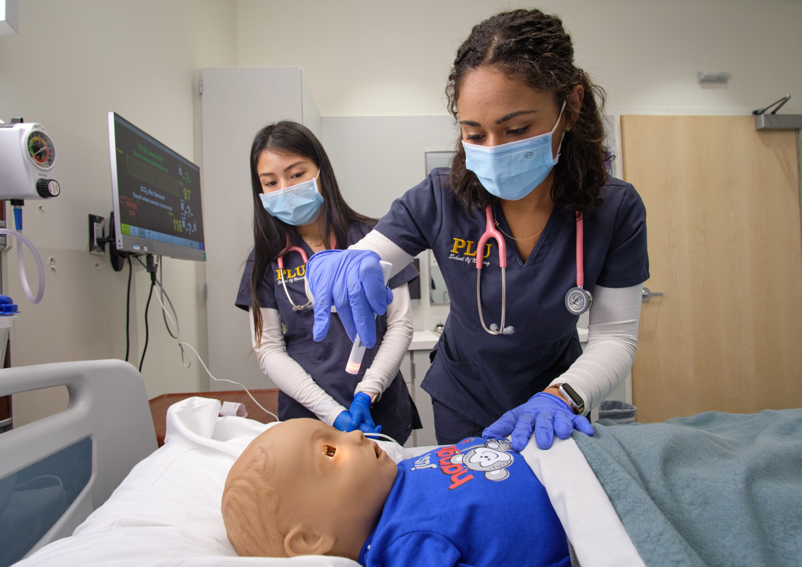 Two PLU nursing students wearing scrubs and facebooks practice a procedure on a child nursing education dummy.