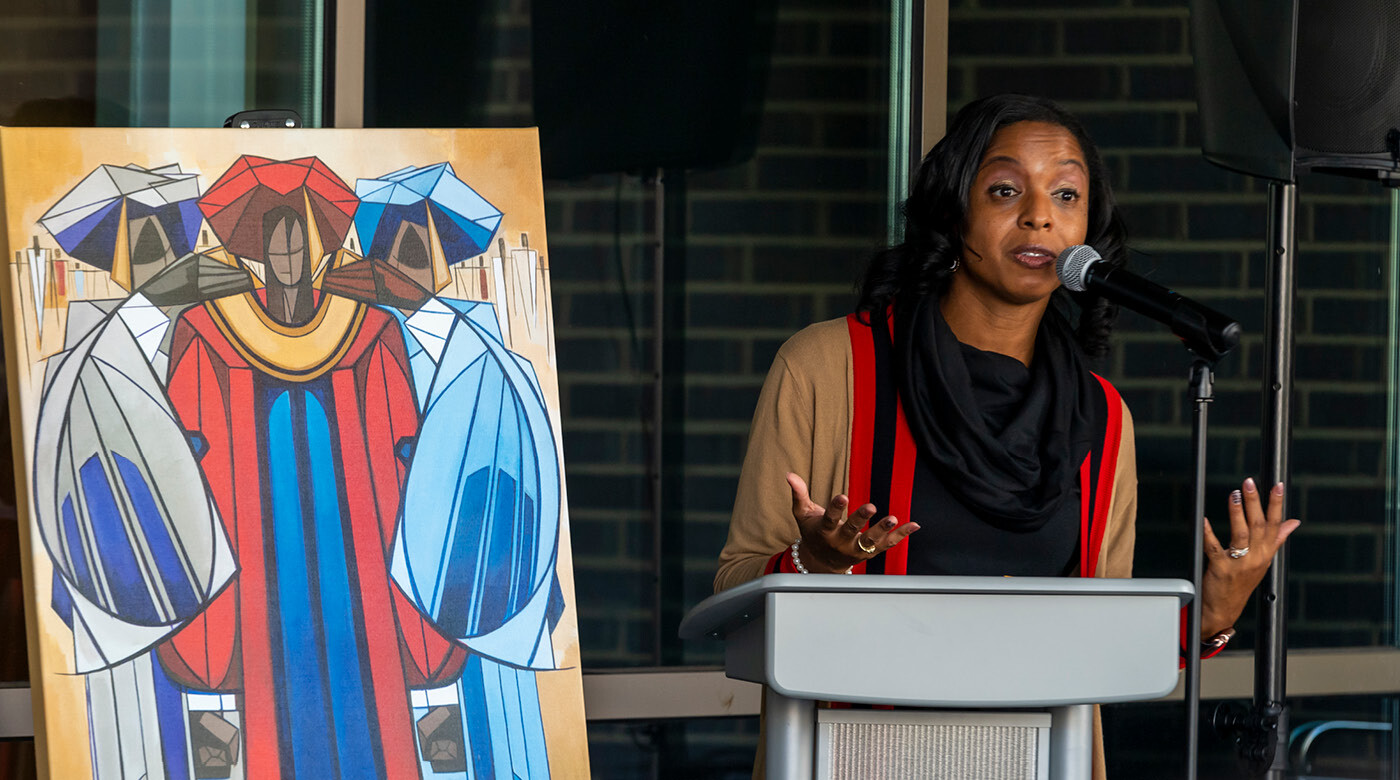 Rihana Mason speaking at a recent event. she's standing at a grey podium with a large piece of art behind her.