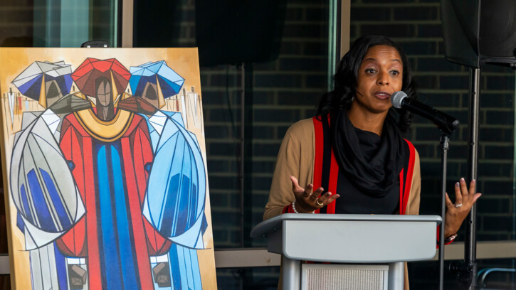 Rihana Mason speaking at a recent event. she's standing at a grey podium with a large piece of art behind her.