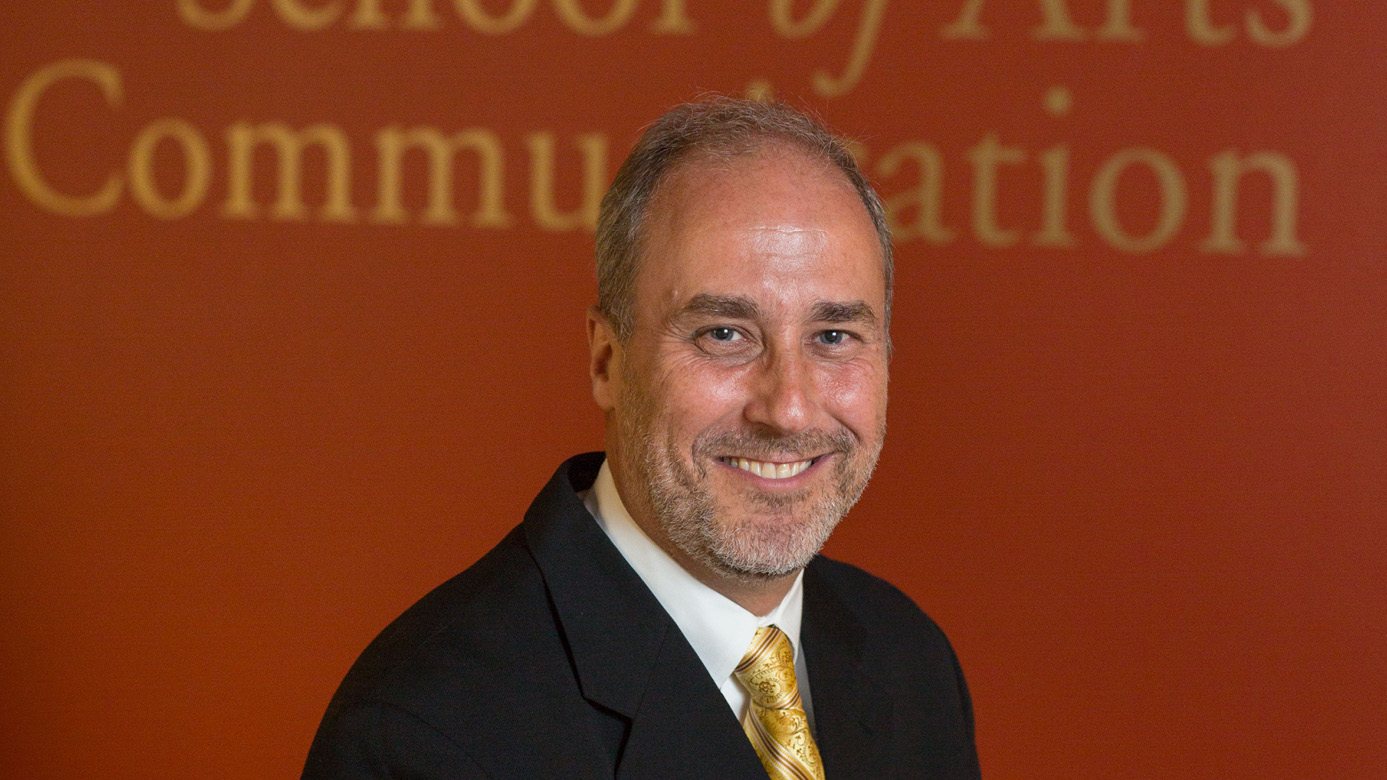 Cameron Bennett smiles while wearing a suit with a red wall behind him