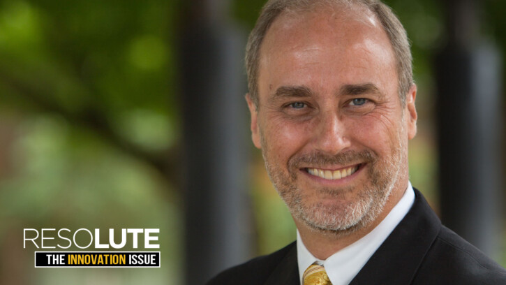 Cameron Bennett smiles in a headshot. He's wearing a suit and tie and there is blurry greenery behind him.