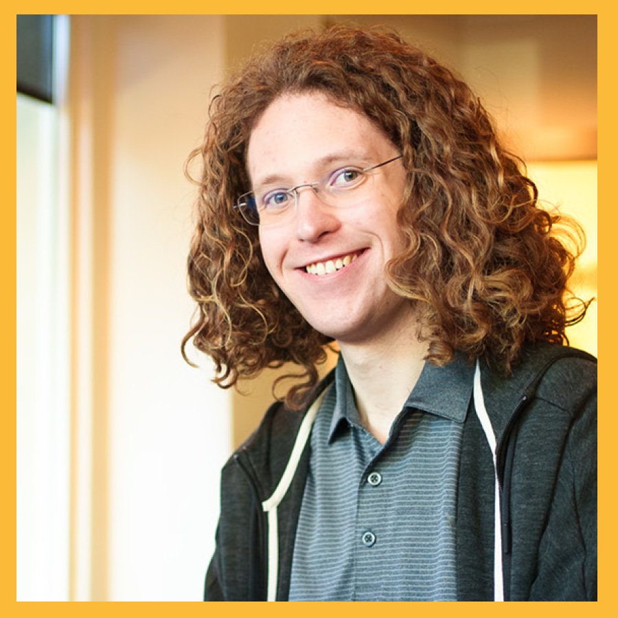 Matthew Conover smiling while sitting in a coffee shop. He has long red hair and is wearing an unzipped black hoodie