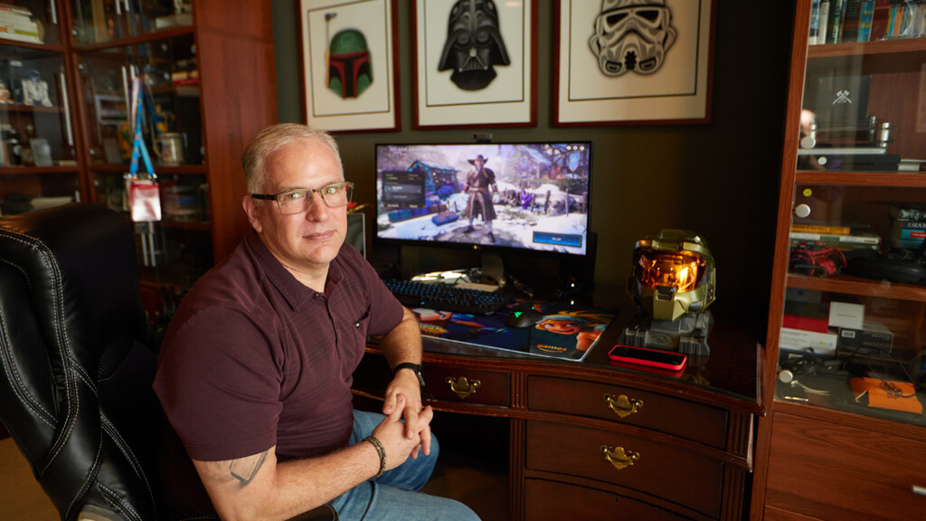 Jon Grande sitting in front of his computer in his own office. His desk and nearby shelves are filled with tokens that remind him of video games he's worked on.