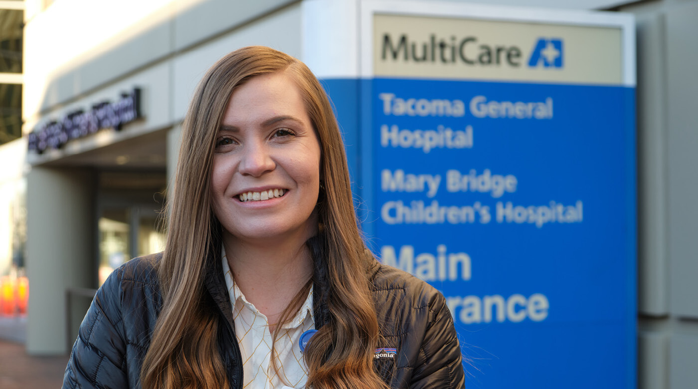 Leah Butters standing outside of Tacoma General Hospital with a blue "MultiCare" sign and logo above her left shoulder.