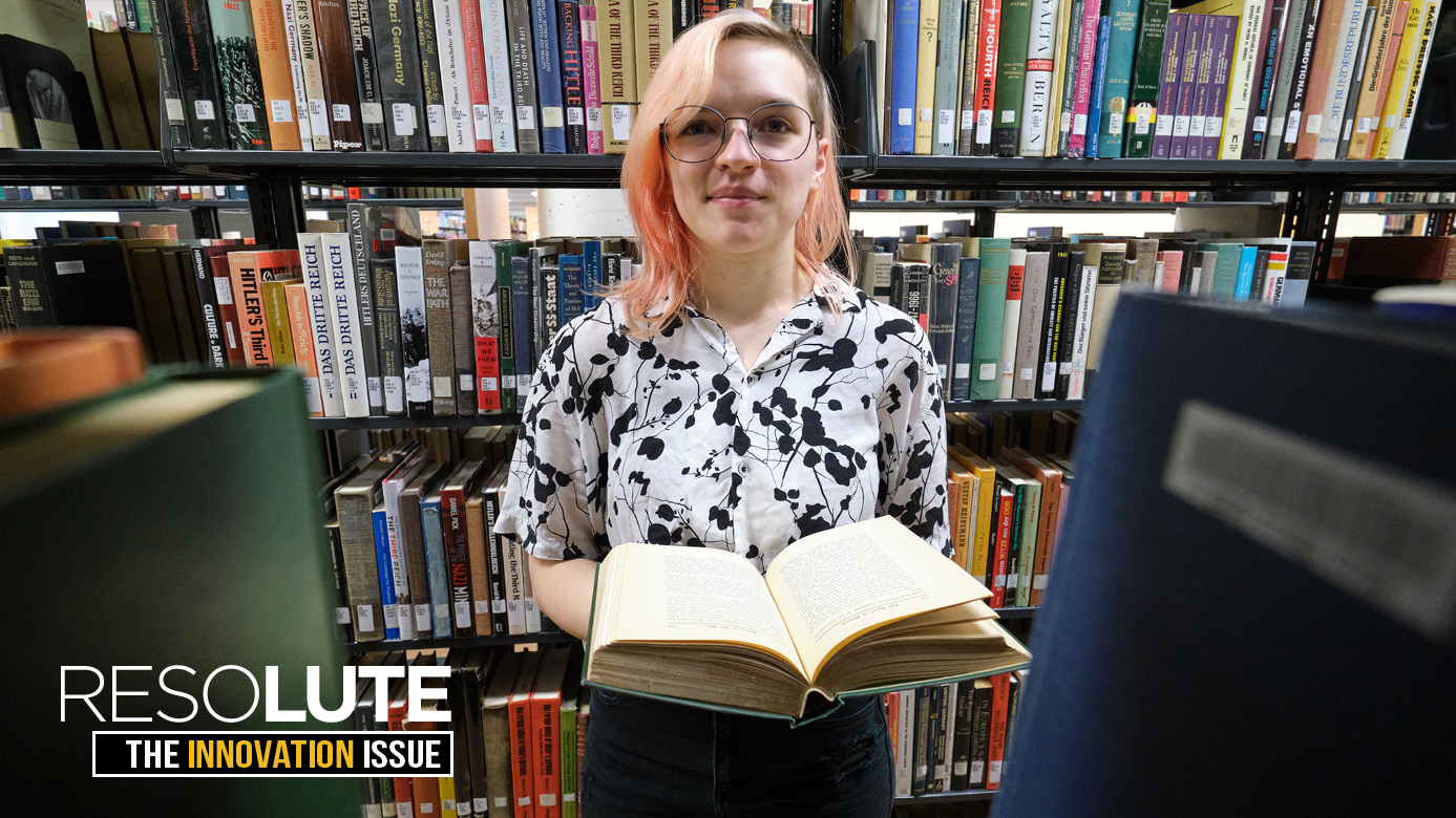 Nicole Query holds a book while standing in an isle of the PLU library surrounded by research books.
