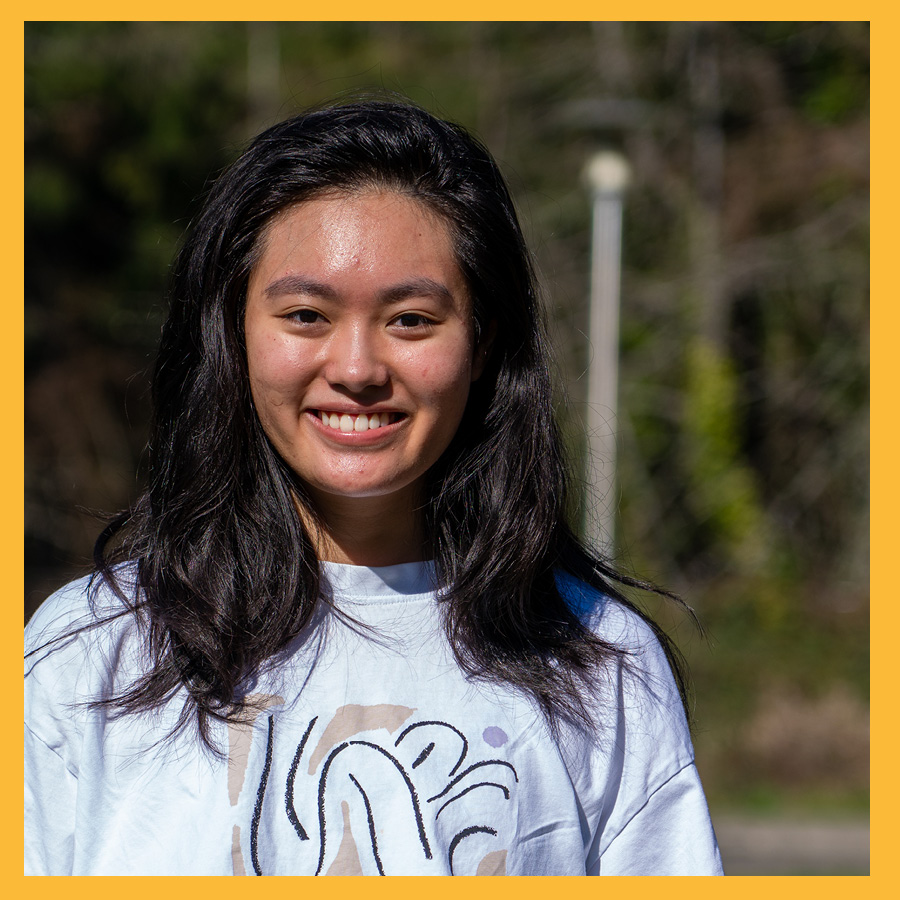 Student Mindy Tieu standing in Foss field on PLU's campus. She's wearing a white crew-neck sweatshirt and smiling.
