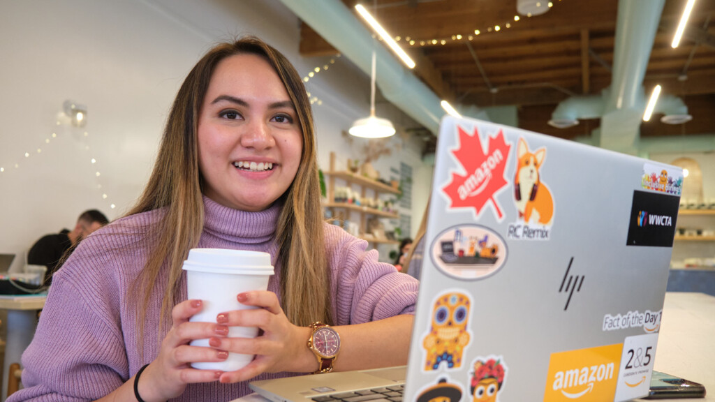 April Rose Nguyen smiles into the camera while sitting in a coffee shop. She's holding a coffee in front of her and has a laptop covered with stickers on the counter in front of her.