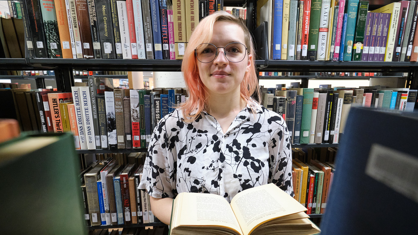 Nicole Query holds a book while standing in an isle of the PLU library surrounded by research books.