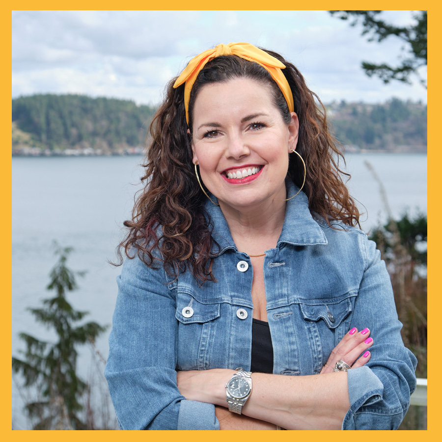 Shelly (Cano) Kurtz wears a denim jacket and yellow bandana in her hair while standing arms folded in front a puget sound beach