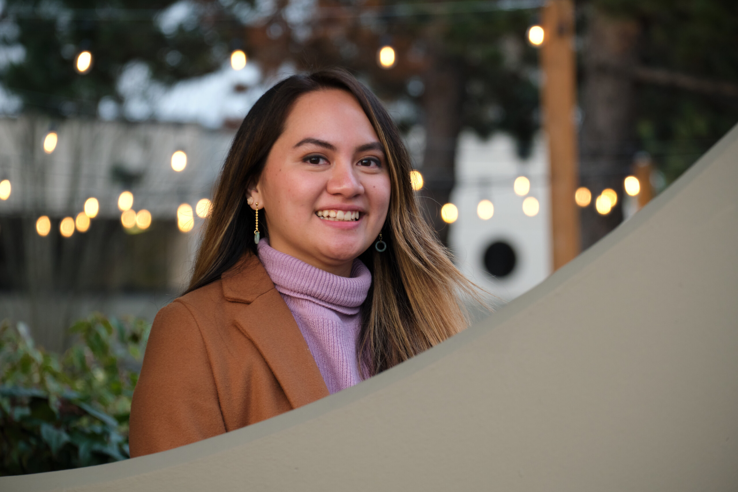April Rose wearing a brown blazer over a pink sweater, smiles into the camera outside with hanging white lights behind her