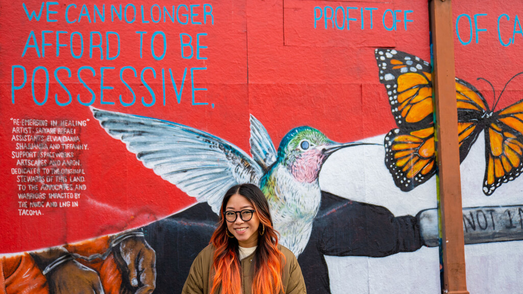 Gines stands in front of a mural of a hummingbird and butterfly that says "we can no longer afford to be posesessive"