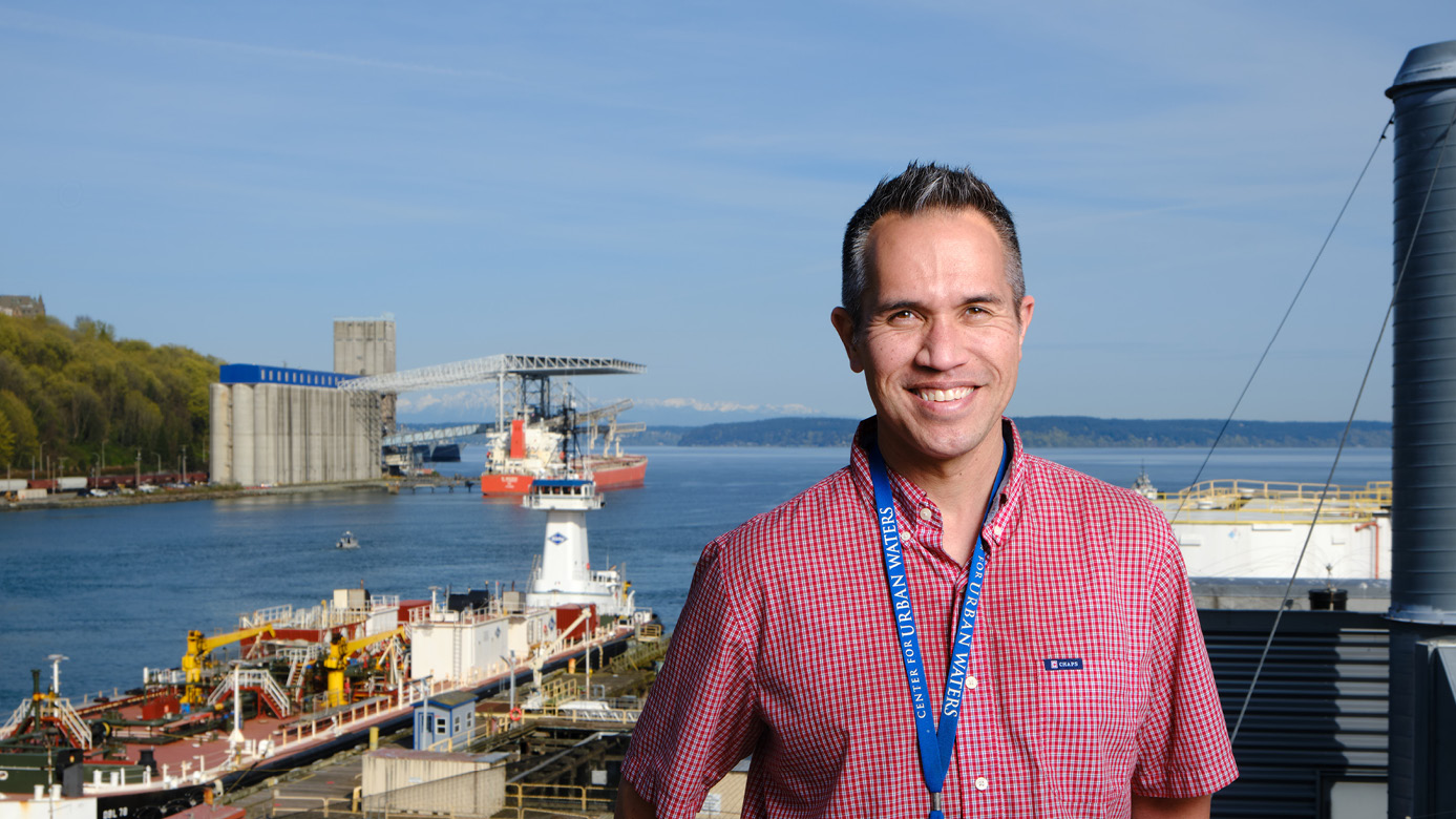 Tom Chontofalsky stands on the deck of the City of Tacoma environmental services offices with the port of tacoma and commencement bay behind him. he's smiling and wearing a short sleeve red button up.