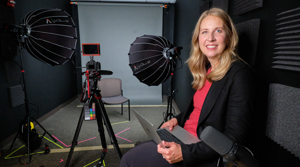 Kristen-lute powered-web PLU alumna Kristen Jaudon '24 poses in a studio setting in front of a photagraphy light set up with a camera on a tripod clearly seen in the background. Jaudon sits with a smile and laptop on her lap.
