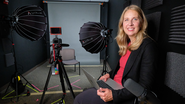 PLU alumna Kristen Jaudon '24 poses in a studio setting in front of a photagraphy light set up with a camera on a tripod clearly seen in the background. Jaudon sits with a smile and laptop on her lap.