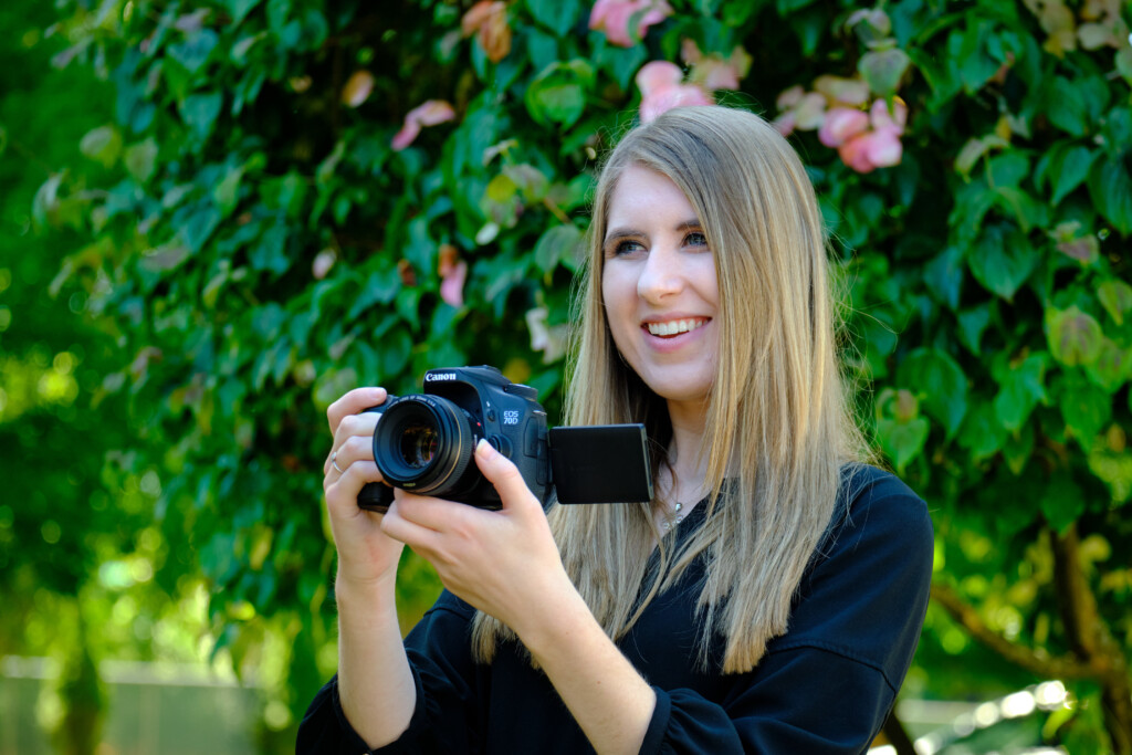 Kate Hall is standing in front of a tree with pink flowers blooming and holding a camera. Kate is smiling while holding the lens of the camera to adjust zoom and smiling.